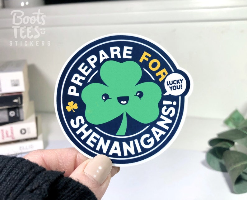 Puede incluir: Una pegatina redonda con fondo azul y borde blanco. La pegatina presenta un tr&eacute;bol verde con una cara sonriente y el texto "Prepare for Shenanigans!" en blanco. Una burbuja de di&aacute;logo amarilla con el texto "Lucky You!" est&aacute; al lado del tr&eacute;bol.