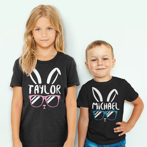 May include: Two children wearing black t-shirts with white bunny ears and sunglasses. The girl's shirt says "TAYLOR" with pink sunglasses. The boy's shirt says "MICHAEL" with blue sunglasses.