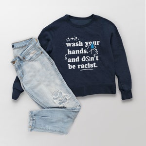 May include: A navy blue sweatshirt with the text "wash your hands, and don't be racist." in white lettering. The sweatshirt is paired with light blue jeans.