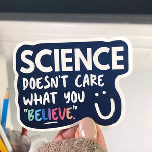 K&ouml;nnte beinhalten: Ein blauer Aufkleber mit wei&szlig;em Text, der "SCIENCE DOESN'T CARE WHAT YOU "BELIEVE"." lautet. Der Text "BELIEVE" ist in Regenbogenfarben.