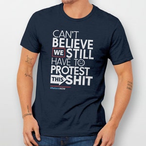 May include: A navy blue t-shirt with white text that reads "Can't Believe We Still Have To Protest This Shit" with a sad face emoji and an arrow pointing to the right. The text also includes the hashtag #ReformNOW.