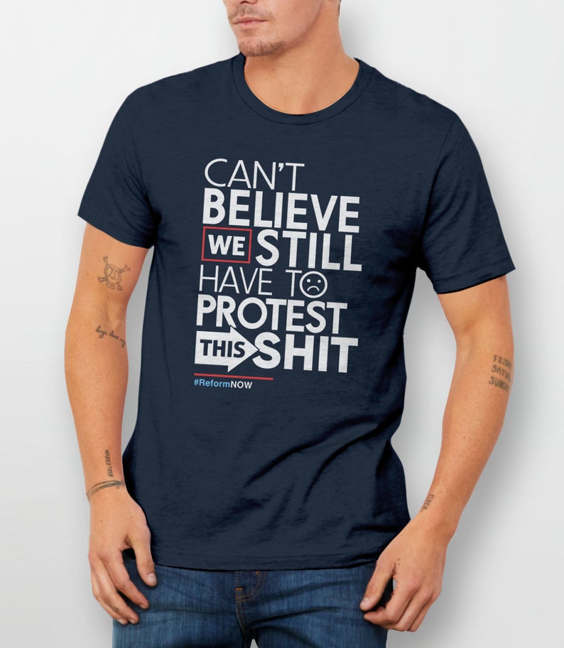 May include: A navy blue t-shirt with white text that reads "Can't Believe We Still Have To Protest This Shit" with a sad face emoji and an arrow pointing to the right. The text also includes the hashtag #ReformNOW.
