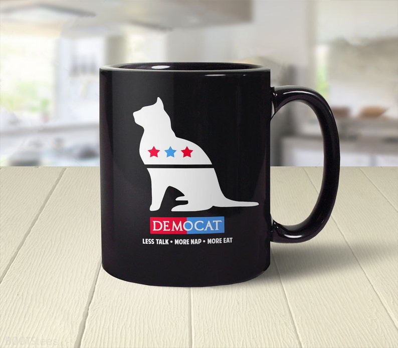 May include: Black coffee mug featuring a white silhouette of a cat with red and blue stars on its chest. The text "DEMOCAT" is below the cat and the text "LESS TALK - MORE NAP - MORE EAT" is below that.