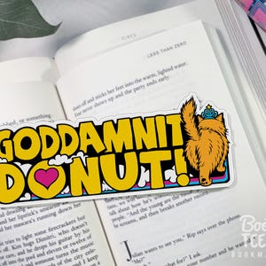 Puede incluir: Una pegatina rectangular con el texto "GODDAMNIT DONUT!" en letras amarillas, un coraz&oacute;n rosa y un gato de dibujos animados con una corona. La pegatina est&aacute; sobre un libro abierto.