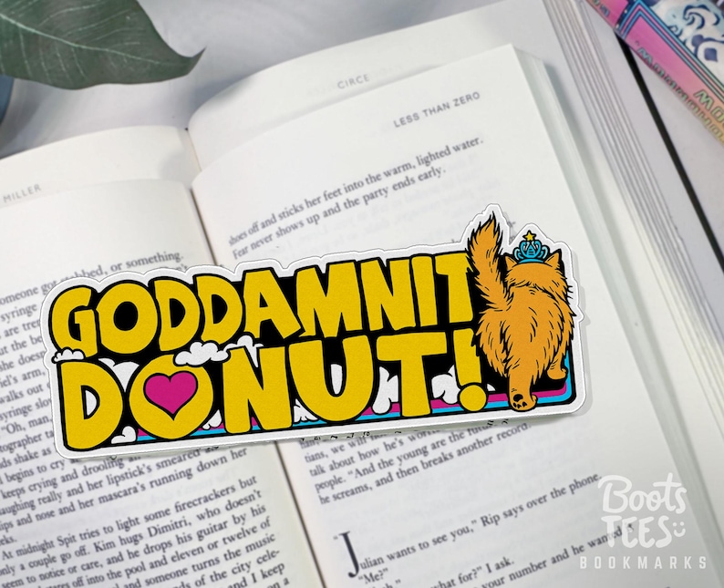 Puede incluir: Una pegatina rectangular con el texto "GODDAMNIT DONUT!" en letras amarillas, un coraz&oacute;n rosa y un gato de dibujos animados con una corona. La pegatina est&aacute; sobre un libro abierto.