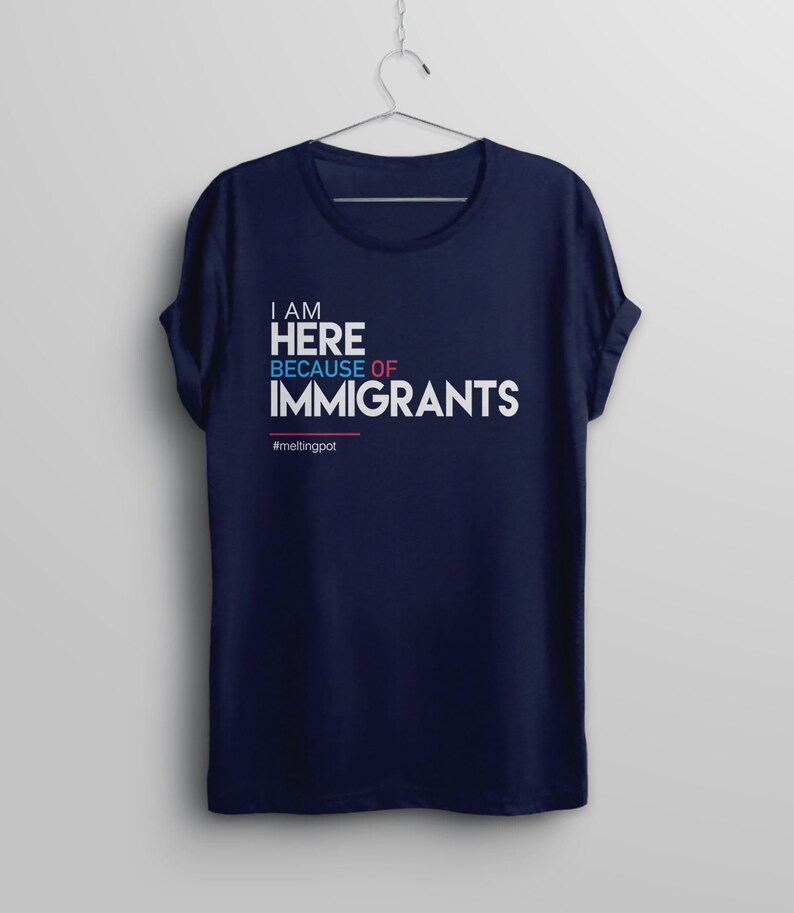 May include: A navy blue t-shirt with the text "I am here because of immigrants #meltingpot" in white, blue, and pink lettering.