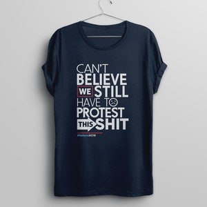 May include: A navy blue t-shirt with white text that reads "Can't Believe We Still Have To Protest This Shit" with a red box around "We" and a red arrow pointing to the right. The text "#ReformNOW" is printed in smaller white letters below the main text.