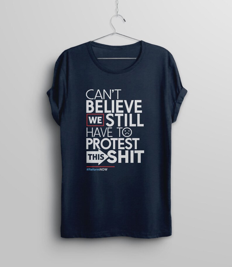 May include: A navy blue t-shirt with white text that reads "Can't Believe We Still Have To Protest This Shit" with a red box around "We" and a red arrow pointing to the right. The text "#ReformNOW" is printed in smaller white letters below the main text.