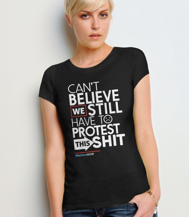 May include: Black t-shirt with white text that reads "Can't Believe We Still Have To Protest This Shit" with a red box around "We" and a red arrow pointing right. The text also includes a sad face emoji and the hashtag #ReformNOW.