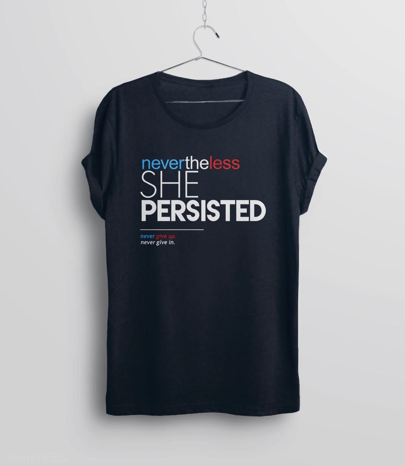 Puede incluir: Una camiseta azul oscuro con el texto "nevertheless SHE PERSISTED" en blanco, rojo y azul. El texto "never give up. never give in." est&aacute; impreso en una fuente m&aacute;s peque&ntilde;a debajo del texto principal.