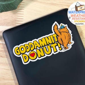 May include: A sticker with the text "GODDAMNIT DONUT!" in yellow with a black outline, a red heart, and an illustration of a fluffy orange cat wearing a crown. The sticker is waterproof and weather-resistant.
