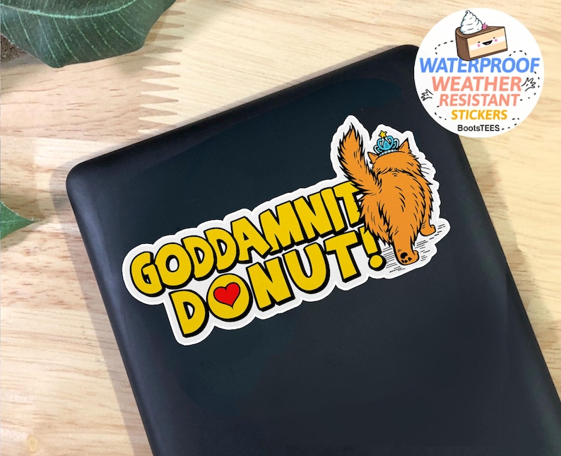 May include: A sticker with the text "GODDAMNIT DONUT!" in yellow with a black outline, a red heart, and an illustration of a fluffy orange cat wearing a crown. The sticker is waterproof and weather-resistant.