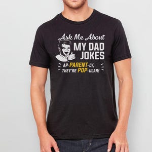 May include: Black t-shirt with a white graphic that says "Ask Me About My Dad Jokes - AP-PARENT-LY, THEY'RE POP-ULAR!" and a cartoon image of a man with a thumbs up.