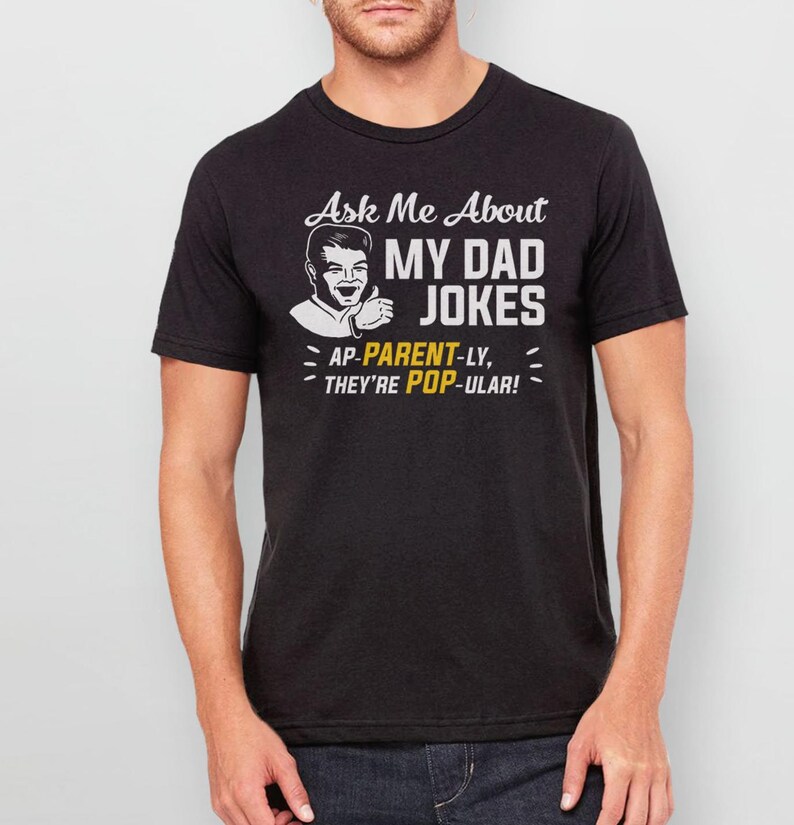 May include: Black t-shirt with a white graphic that says "Ask Me About My Dad Jokes - AP-PARENT-LY, THEY'RE POP-ULAR!" and a cartoon image of a man with a thumbs up.