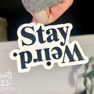 Op de afbeelding: Witte sticker met zwarte tekst die "Stay Weird" zegt.