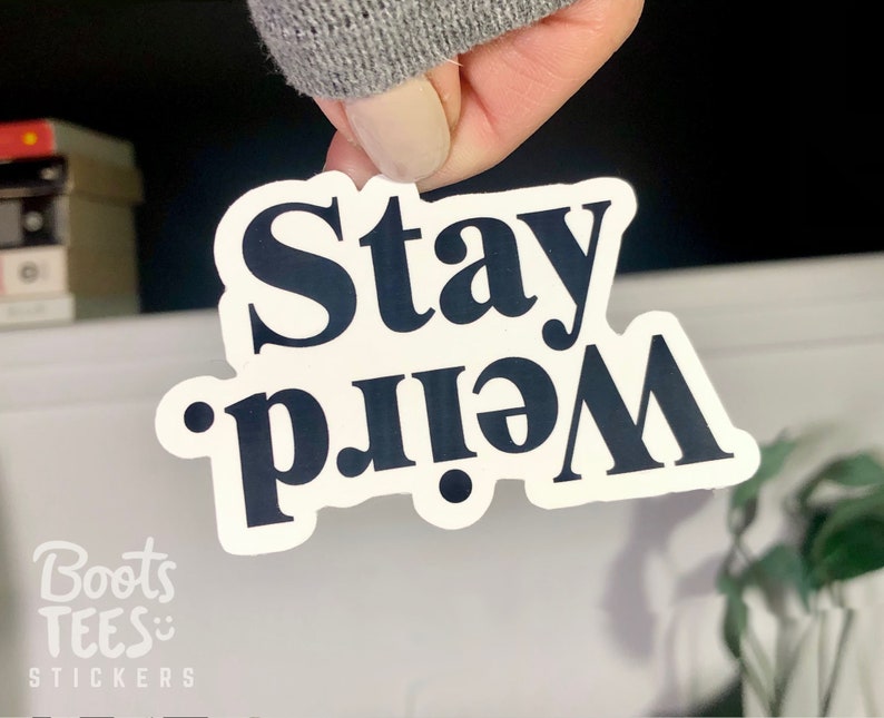Op de afbeelding: Witte sticker met zwarte tekst die "Stay Weird" zegt.