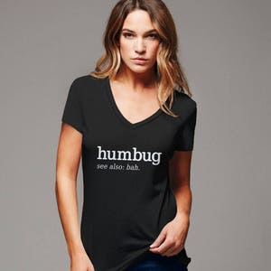 K&ouml;nnte beinhalten: Schwarzes V-Ausschnitt-T-Shirt mit dem wei&szlig;en Wort "humbug" und "see also: bah." Das T-Shirt hat kurze &Auml;rmel. Das Model tr&auml;gt das T-Shirt. Das T-Shirt ist aus weichem Material.