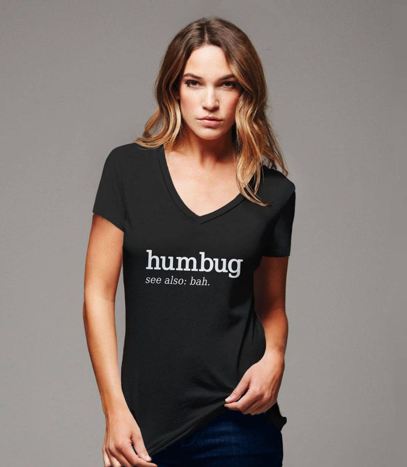 K&ouml;nnte beinhalten: Schwarzes V-Ausschnitt-T-Shirt mit dem wei&szlig;en Wort "humbug" und "see also: bah." Das T-Shirt hat kurze &Auml;rmel. Das Model tr&auml;gt das T-Shirt. Das T-Shirt ist aus weichem Material.
