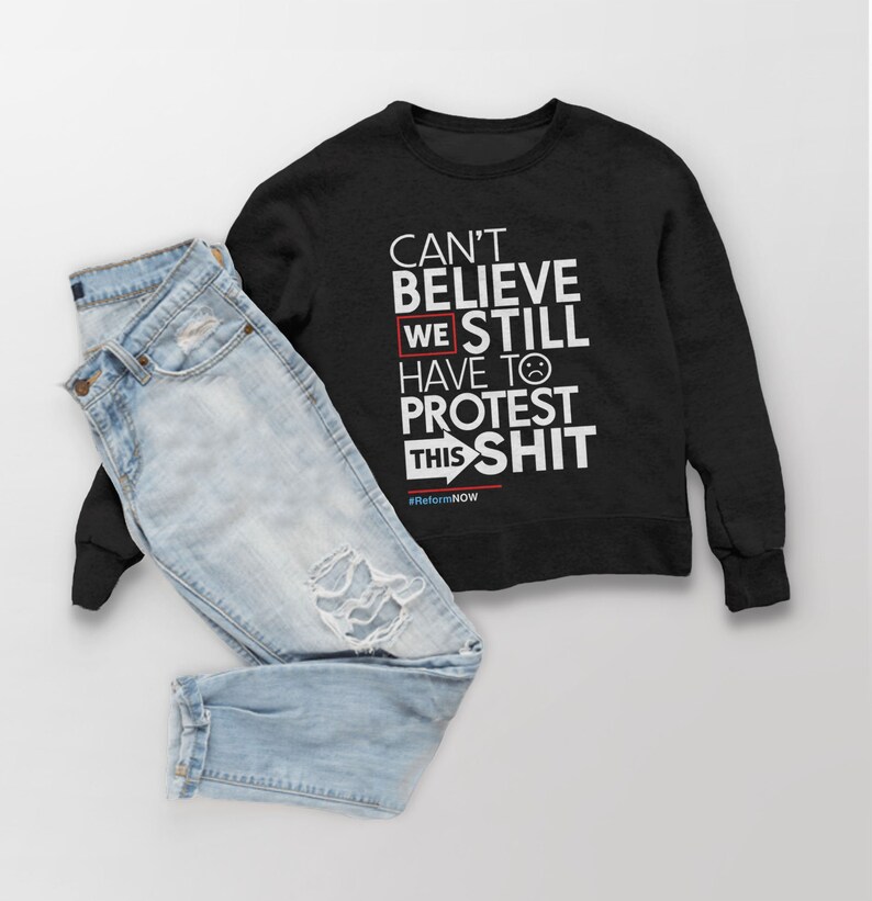 May include: A black sweatshirt with white text that reads "CAN'T BELIEVE WE STILL HAVE TO PROTEST THIS SHIT" and the hashtag #ReformNOW. Light blue denim jeans with distressed details are next to the sweatshirt.