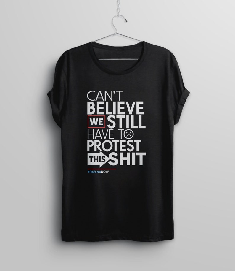 May include: Black t-shirt with white text that reads "Can't Believe We Still Have To Protest This Shit" with a red rectangle around "We" and a red arrow pointing to the right. The text "#ReformNOW" is in smaller white letters below the main text.