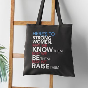 May include: Black tote bag with white text that reads "Here's to strong women, may we know them, may we be them, may we raise them."