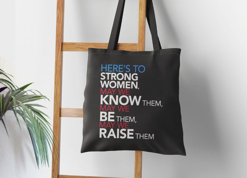 May include: Black tote bag with white text that reads "Here's to strong women, may we know them, may we be them, may we raise them."