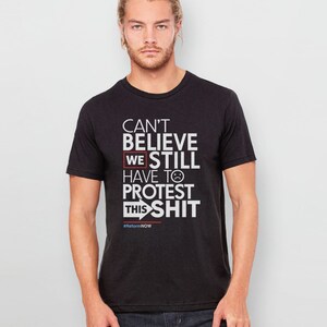 May include: Black t-shirt with white text that reads "Can't Believe We Still Have To Protest This Shit" with a red box around "We" and a sad face emoji. The text also includes the hashtag #ReformNOW.