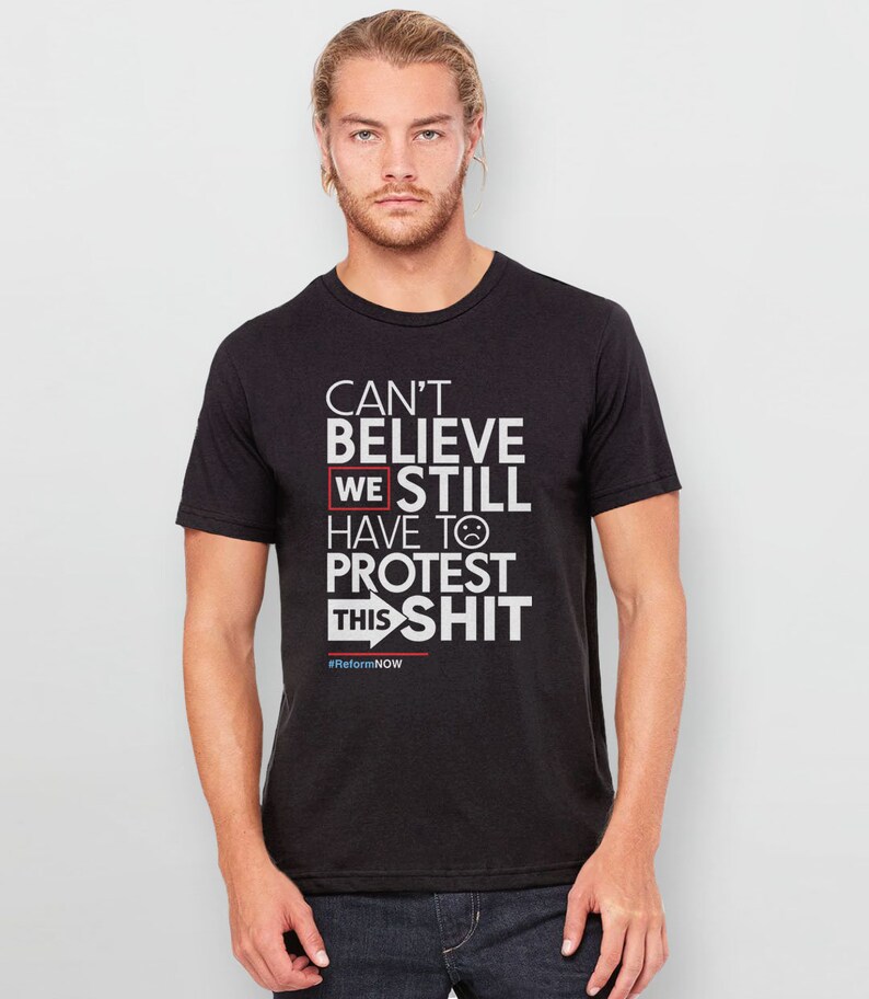 May include: Black t-shirt with white text that reads "Can't Believe We Still Have To Protest This Shit" with a red box around "We" and a sad face emoji. The text also includes the hashtag #ReformNOW.