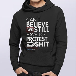 May include: Black hooded sweatshirt with white text: "CAN'T BELIEVE WE STILL HAVE TO PROTEST THIS SHIT." The word "WE" is highlighted in a red box. The text includes a sad face emoji and the hashtag #ReformNOW.