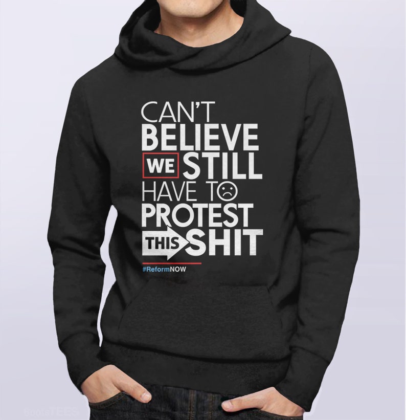 May include: Black hooded sweatshirt with white text: "CAN'T BELIEVE WE STILL HAVE TO PROTEST THIS SHIT." The word "WE" is highlighted in a red box. The text includes a sad face emoji and the hashtag #ReformNOW.