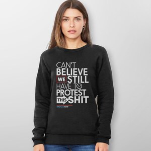 May include: Black sweatshirt with white text that reads "CAN'T BELIEVE WE STILL HAVE TO PROTEST THIS SHIT" with a sad face emoji. The text "#ReformNOW" is at the bottom. The sweatshirt is worn by a person.