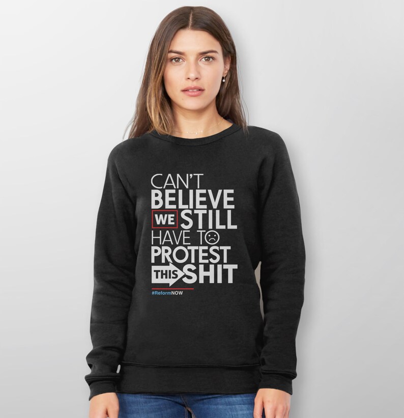 May include: Black sweatshirt with white text that reads "CAN'T BELIEVE WE STILL HAVE TO PROTEST THIS SHIT" with a sad face emoji. The text "#ReformNOW" is at the bottom. The sweatshirt is worn by a person.