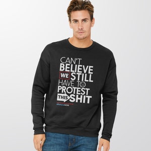 May include: Black sweatshirt with white text that reads "CAN'T BELIEVE WE STILL HAVE TO PROTEST THIS SHIT" with a sad face emoji. The text "WE" is highlighted in a red box. The hashtag "#ReformNOW" is at the bottom.