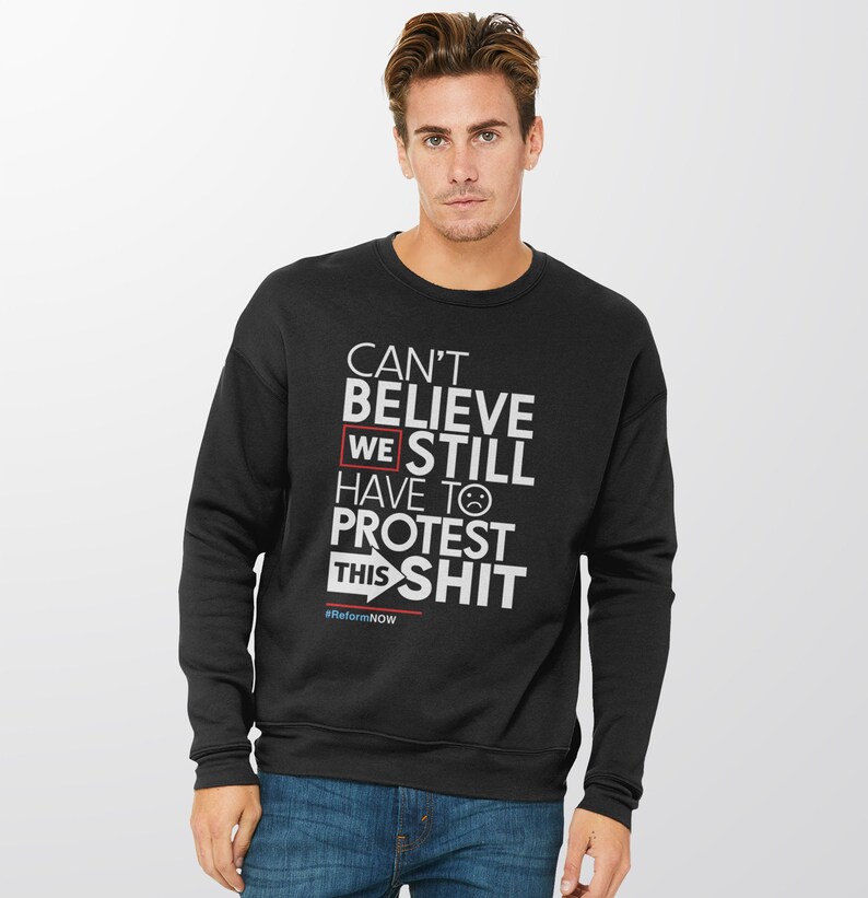May include: Black sweatshirt with white text that reads "CAN'T BELIEVE WE STILL HAVE TO PROTEST THIS SHIT" with a sad face emoji. The text "WE" is highlighted in a red box. The hashtag "#ReformNOW" is at the bottom.