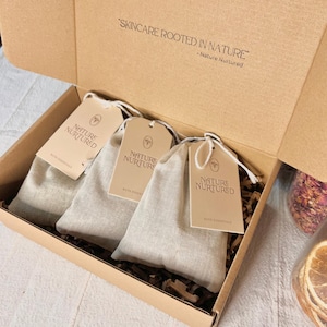 May include: A cardboard box holding three linen pouches with tags. The tags say "NATURE NURTURED" and "BATH ESSENTIALS". The box lid features the text "SKINCARE ROOTED IN NATURE - Nature Nurtured".