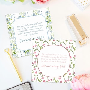 May include: Two square cards with floral borders and scriptural text. One card has blue flowers and the text "Proverbs 31:25,28". The other card has pink flowers and the text "Deuteronomy 31:8". A gold pen and stapler are also visible.