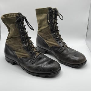 May include: A pair of high-top combat boots featuring black leather lower sections and olive green canvas uppers. The boots include black laces, metal eyelets, and thick black rubber lug soles, providing a sturdy construction for these tactical-style boots.