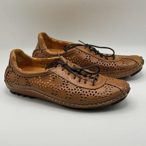 May include: A pair of tan leather lace-up shoes featuring intricate laser-cut floral patterns across the sides and toe box. The shoes have dark brown laces, visible light-colored stitching along the sole edges, and a low-profile rubber outsole.