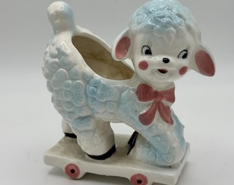 May include: A vintage ceramic lamb planter in white and light blue, with pink accents on the ears, cheeks, and a bow. The lamb is mounted on a white base with pink wheels. The planter has a hollowed-out area for holding small plants or decorative items.