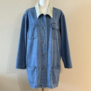 May include: A long-sleeve blue denim jacket featuring a light beige corduroy collar, a front button-down closure with metal buttons, two chest pockets with button flaps, and two large open patch pockets at the lower front of the garment.