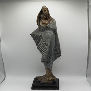 May include: A bronze-toned sculpture depicting two figures, one larger and one smaller, wrapped in a textured gray shawl with geometric patterns. The figures stand on a dark rectangular base, with the larger figure embracing the smaller one.