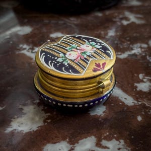 Puede incluir: Pequeña caja de baratijas antigua con marco de metal dorado. La tapa es azul marino con un diseño floral y detalles dorados. La caja es redonda con tapa abisagrada y cierre decorativo.