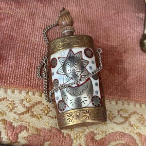 May include: A small, ornate perfume bottle with a silver sun and moon design on a white background. The bottle has a wooden stopper and is trimmed with gold-colored metal. A delicate chain is attached for carrying.