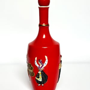 Può includere: Una vivace bottiglia in ceramica rossa con un corpo arrotondato e un lungo collo, sormontata da un tappo rosso. La bottiglia presenta figure in rilievo di persone in abiti tradizionali. Una fascia dorata adorna il collo.