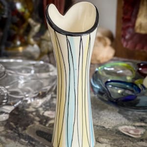 May include: A cream-coloured ceramic vase with a heart-shaped opening. The vase is decorated with vertical black, light blue, and yellow lines. The rim is outlined with a black band. The vase is a decorative item.