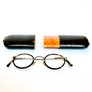 Peut inclure: Lunettes rondes vintage avec monture et branches à motif écaille de tortue. Les lunettes sont placées devant un étui à lunettes en cuir noir et marron, ouvert pour révéler un intérieur orange.