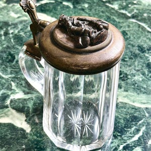 Può includere: Un boccale di birra in vetro trasparente con coperchio e manico decorativi in metallo. Il coperchio presenta un design a rilievo dettagliato. Il vetro ha un motivo a stella sul fondo. Il boccale è su una superficie di marmo verde.