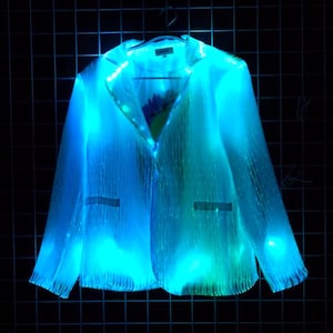 May include: A glowing, futuristic blazer in shades of blue and green hangs against a dark background. The jacket has a classic design with lapels, pockets, and a button closure, illuminated with fiber optic lights.