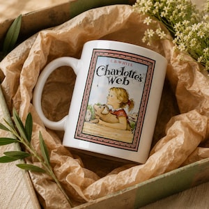 May include: A white ceramic mug featuring the book cover illustration of Charlotte's Web by E.B. White. The graphic shows a young girl holding a piglet, framed by a pink border. The text reads E.B. White, Charlotte's Web, and Illustrated by Garth Williams.