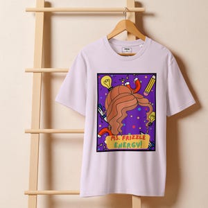Puede incluir: Camiseta de color lila claro con un gráfico colorido. El diseño incluye un peinado de dibujos animados, bombillas, un lápiz y un imán. El texto "MS. FRIZZLE ENERGY!" está en una pancarta amarilla.